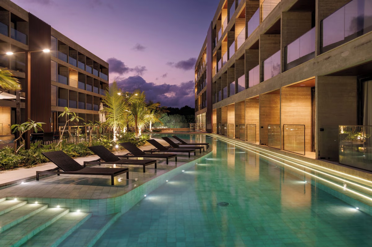 An inviting outdoor pool is highlighted, surrounded by modern buildings. The space is adorned with lounge chairs and features soft lighting along the pool's edge, creating a serene atmosphere as twilight sets in. Lush greenery adds to the tranquil ambiance.