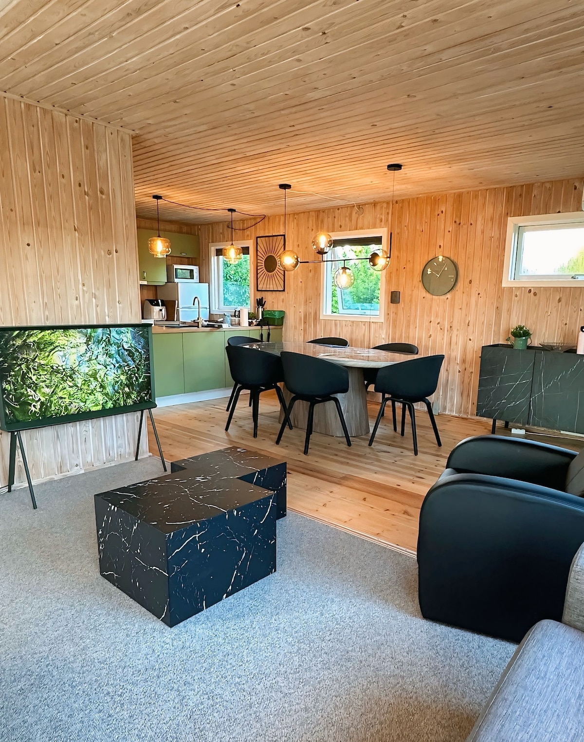 A light-filled living area features wooden walls and ceiling, creating a warm ambiance. A stylish dining table with black chairs is located adjacent to a modern kitchen. A sleek coffee table with a dark finish complements the seating area, which includes two comfortable armchairs.