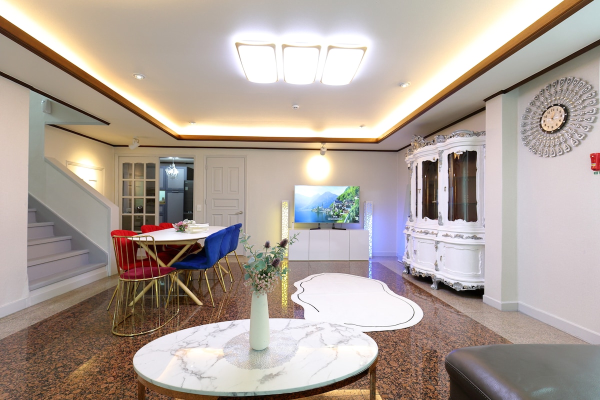 An inviting living area is visible, featuring a spacious marble coffee table at the center. A dining table is set with colorful chairs, while a contemporary white cabinet holds decorative items. A TV unit showcases greenery, and warm lighting creates a bright atmosphere.