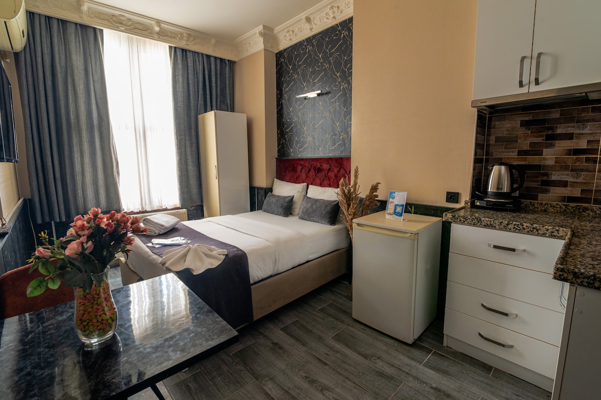 A well-lit room features a spacious bed adorned with decorative pillows and a soft blanket. A small kitchenette is present, equipped with a kettle and a compact refrigerator. A table with a floral arrangement adds a touch of greenery, while large windows allow natural light to fill the space.
