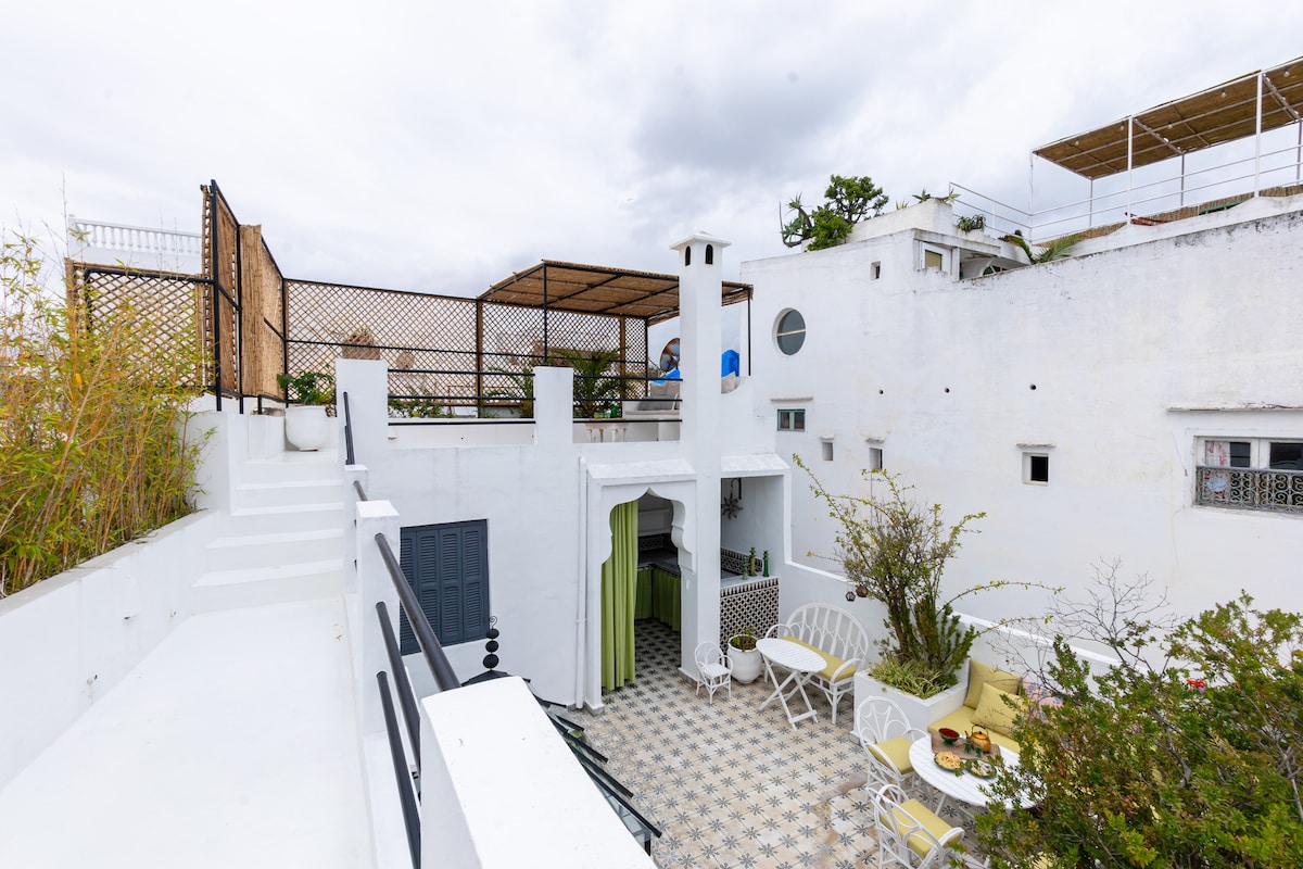 A tranquil courtyard is displayed, featuring a patterned tile floor and white walls. A seating area with round chairs is framed by greenery. A staircase leads to a rooftop terrace, while a small kitchen area is partially visible, enhancing the inviting outdoor space.