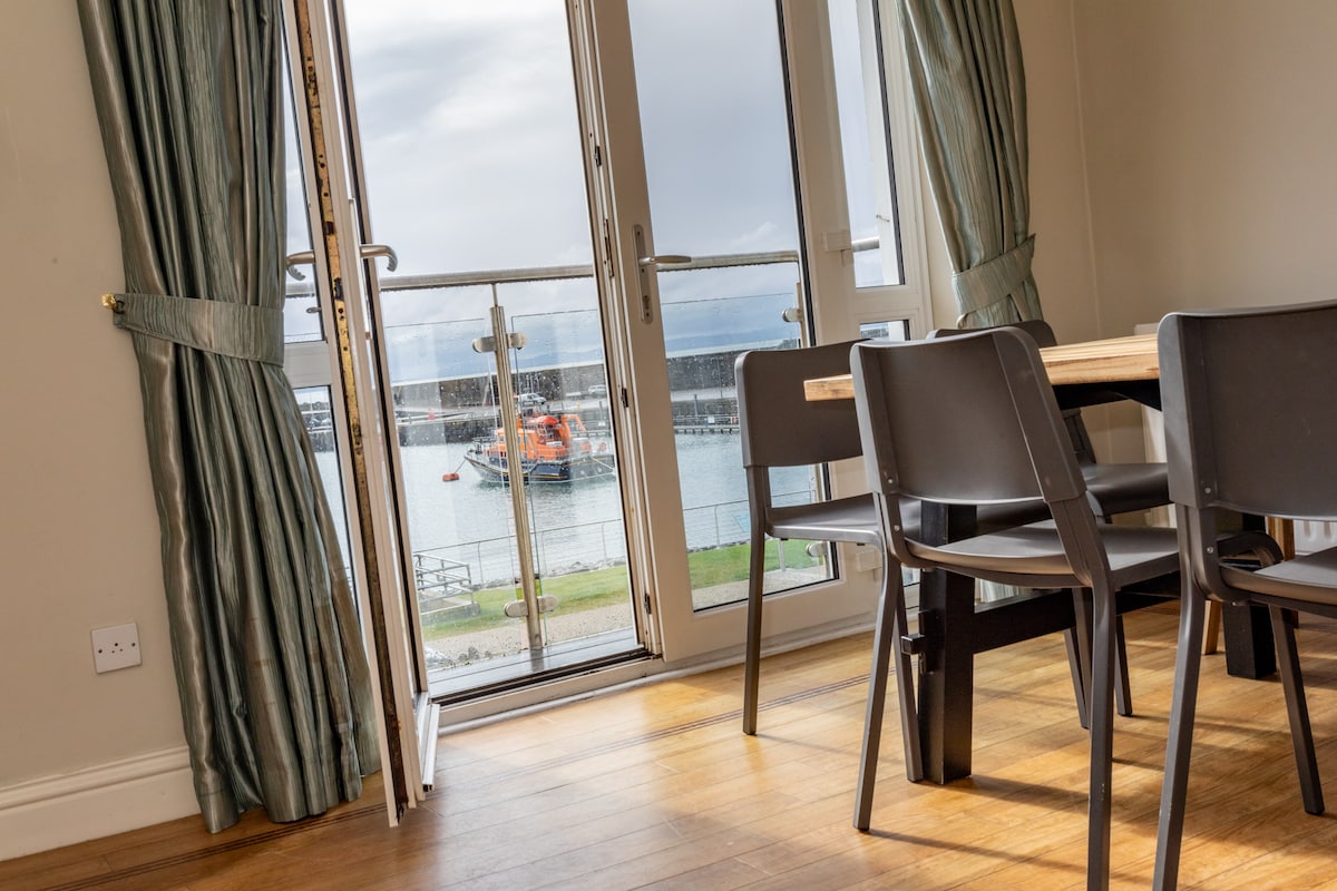 A dining area features a wooden table surrounded by black chairs, with expansive glass doors opening to a view of the harbour. Natural light enters through the doors, and curtains in a muted shade frame the scene.