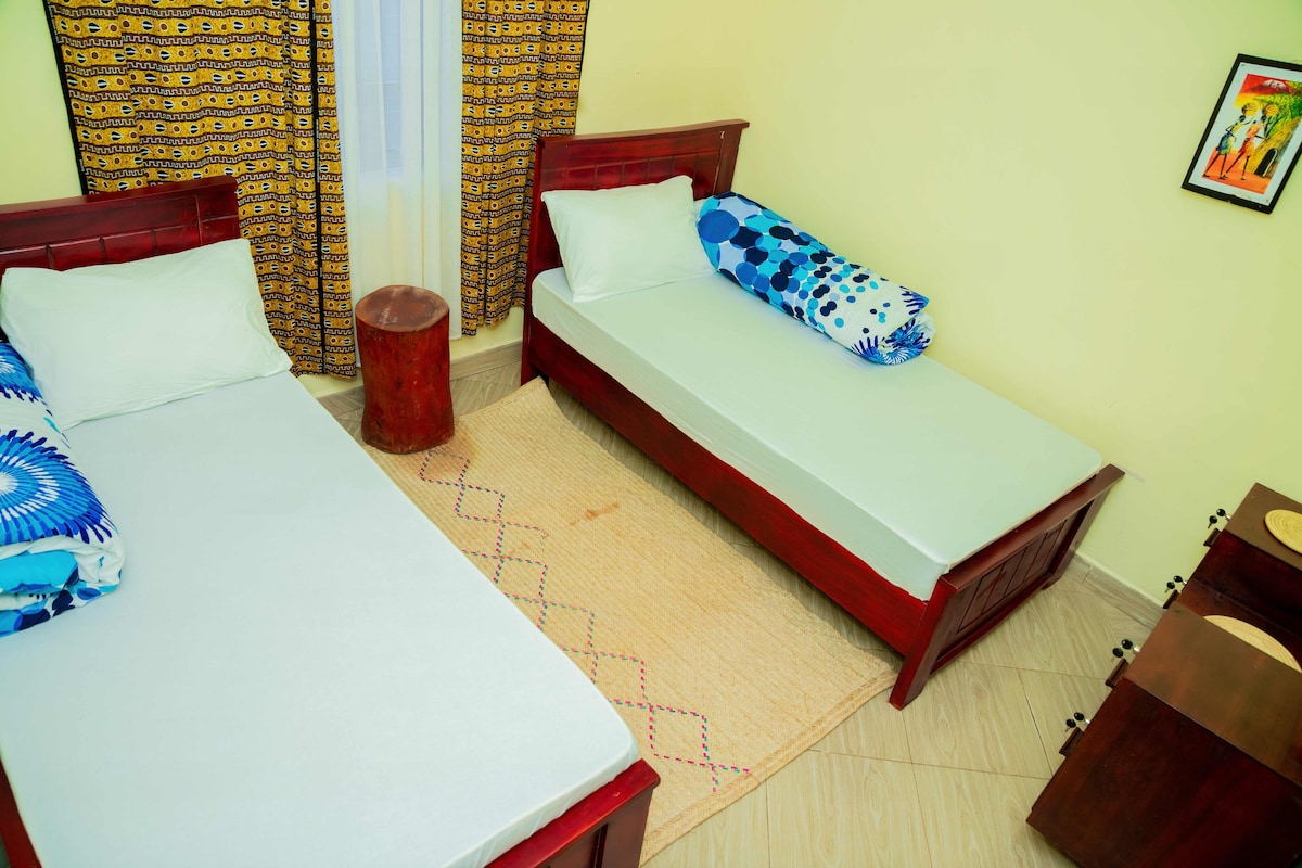 A cozy sleeping area contains two individual beds adorned with simple linens and vibrant throw pillows. A round wooden table stands between them, complimented by a light-colored rug. The room features patterned curtains that frame the window, adding a colorful touch to the space.