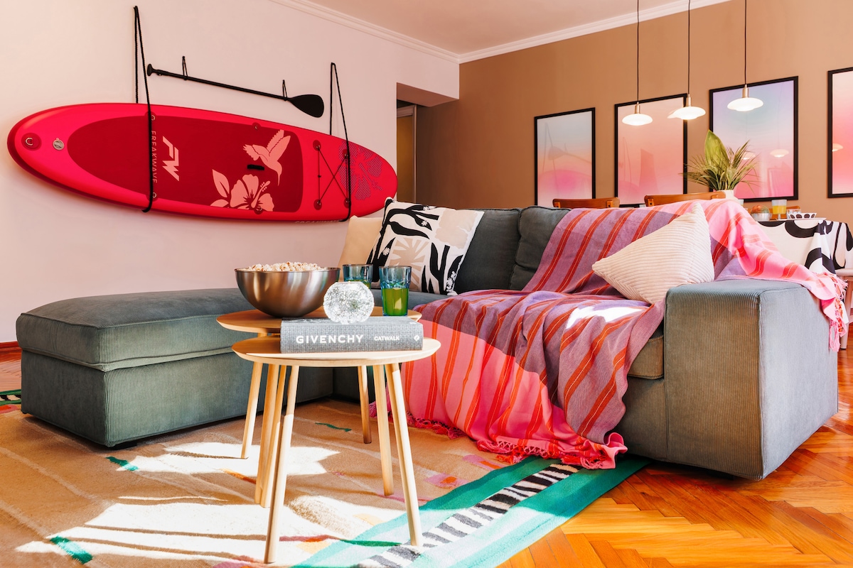 A spacious living room is showcased with a large sectional couch, adorned with a colorful blanket. A decorative coffee table displays stylish books and a decorative orb. A bright paddleboard is mounted on the wall, complementing the vibrant artwork and the warm wooden floors.