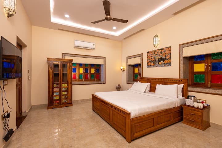 Bedroom 4 – Rang Bhawan: Vibrant and inviting, with a large wooden bed, white bedding, stained glass windows, AC, TV, and cozy seating. Floral artwork and warm lighting add charm to this colorful, heritage-inspired retreat