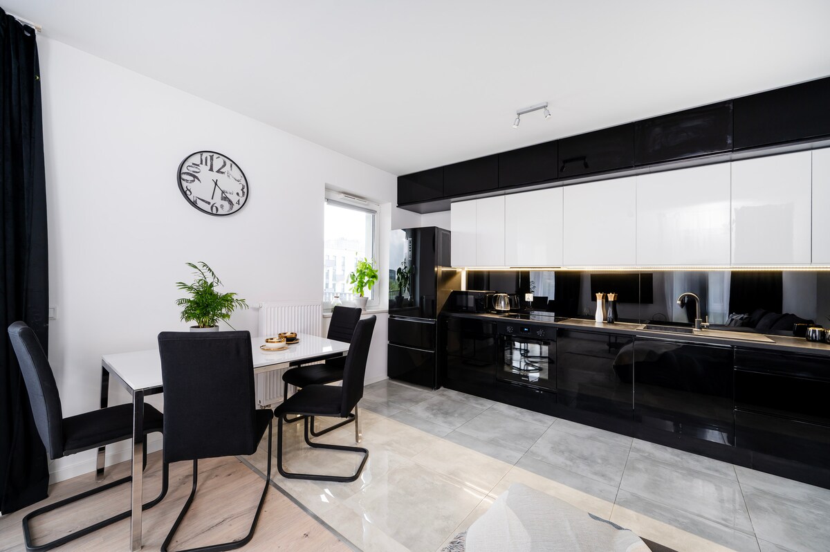 A modern kitchen is showcased with a sleek black and white cabinetry design. A dining table with four chairs is visible, complemented by a stylish clock on the wall. Natural light is provided by a window, enhancing the open and functional layout.