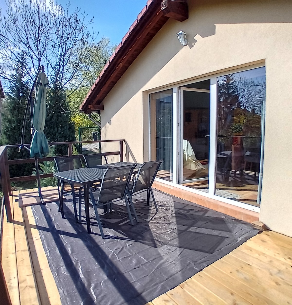 A wooden terrace is featured, equipped with a table and chairs for outdoor dining. A green umbrella offers shade. The adjacent glass doors provide access to the interior, surrounded by greenery that enhances the peaceful ambiance.