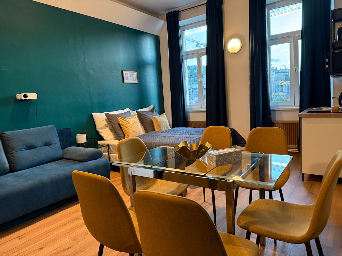 The open-plan living area features a glass dining table surrounded by yellow chairs. A spacious bed with neutral-toned bedding occupies one corner, while a dark blue sofa is positioned nearby. Large windows allow natural light to fill the room, adorned with navy curtains.