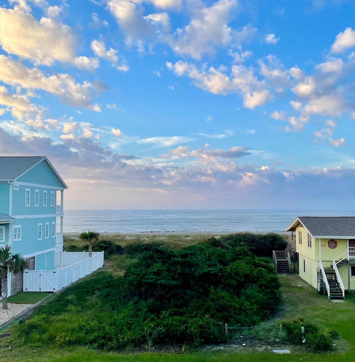 A view of the expansive ocean from the property, framed by lush greenery and neighboring coastal homes. The sky is filled with soft clouds in various shades, reflecting the light of early evening. The tranquil scene captures the inviting atmosphere of a beach retreat.