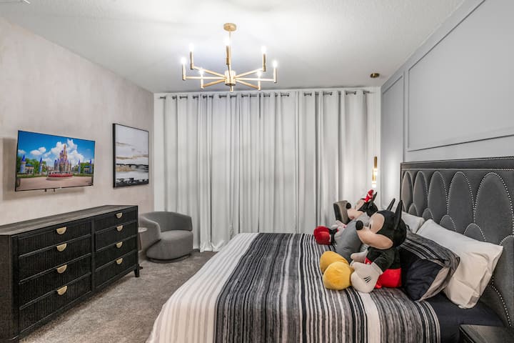 Plongez dans un havre de paix chic : murs gris lambrissés, tête de lit en velours festonné, couches neutres et pendentifs en laiton sous un lustre moderne, où l'ambiance sereine de l'hôtel rencontre une touche ludique de Mickey et Minnie pour des nuits de rêve sans effort.            