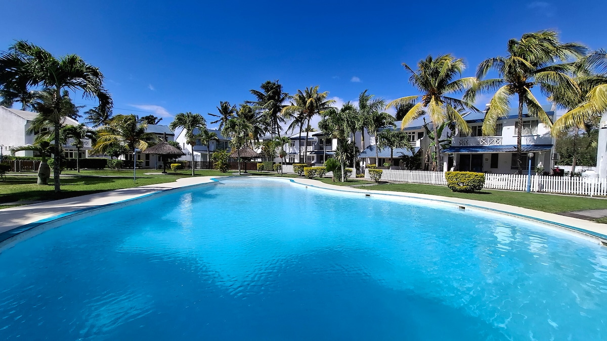 A spacious outdoor pool is situated amidst well-maintained gardens with palm trees swaying gently in the breeze. The clear blue water of the pool reflects the bright sky, inviting relaxation. The surrounding area includes a view of nearby residences and open green spaces.