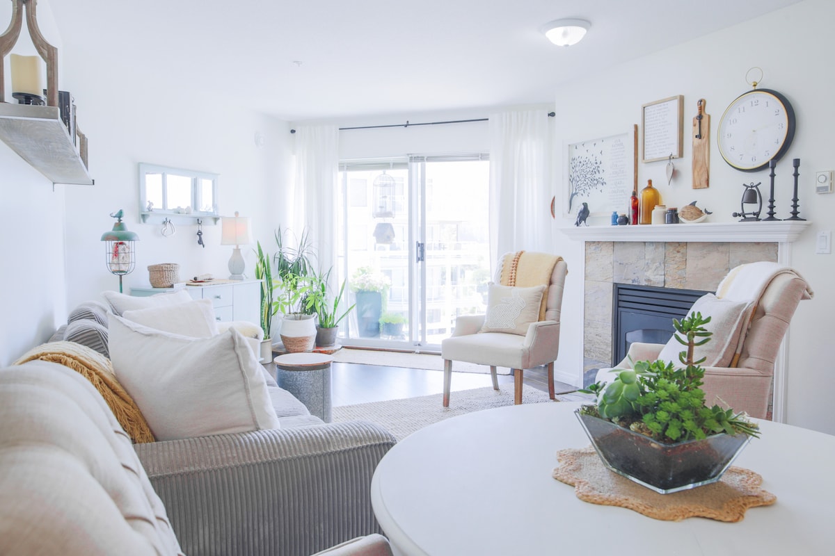 The living room is designed with a comfortable sofa and an armchair, accented by soft throw pillows. A natural stone fireplace serves as a focal point, while large glass doors allow for abundant natural light and access to outdoor views. Decorative plants add a touch of greenery.