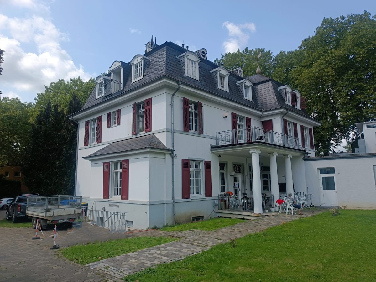 The villa features a grand architectural design with red shutters and a white facade. A spacious patio with seating is visible, alongside a grassy area, enhancing the outdoor environment. A vehicle is parked nearby, with additional outdoor seating arranged in the vicinity.