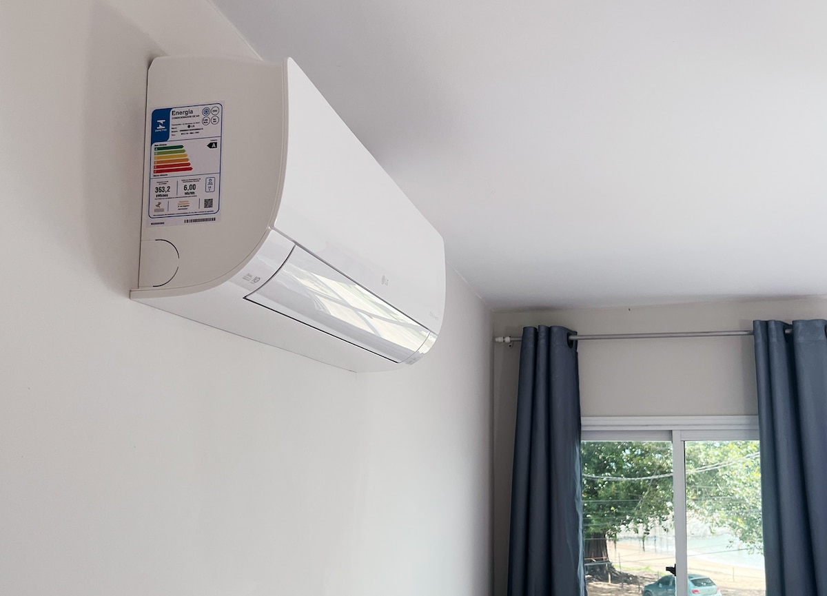 An air conditioning unit is mounted on a white wall, featuring a clear energy rating label. Two long, light gray curtains frame a window that offers a view of greenery outside.