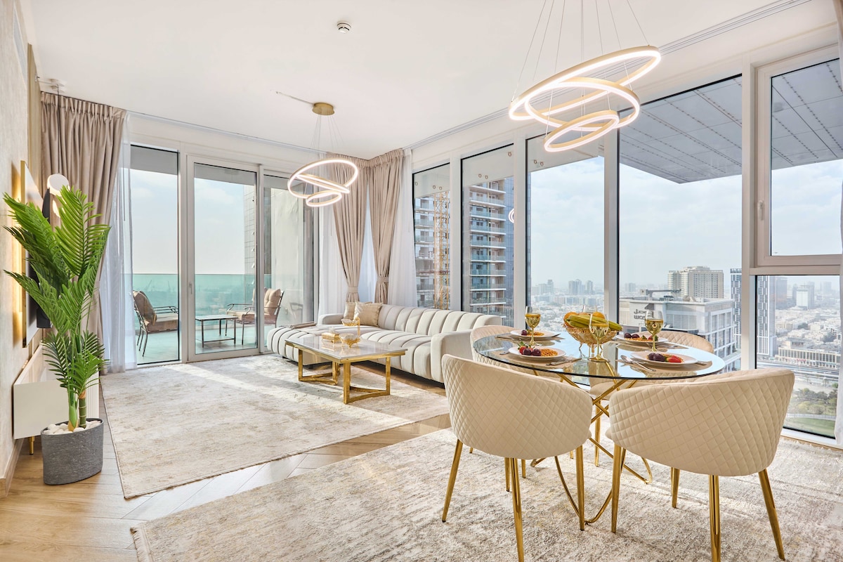A contemporary living and dining area is presented with large windows offering views of the city skyline. A stylish sectional sofa and two elegant dining chairs surround a circular table, while modern light fixtures enhance the space. A potted plant adds a touch of greenery.