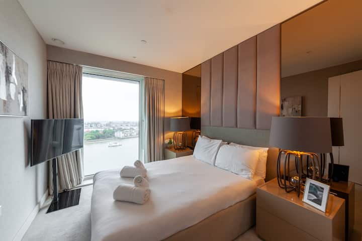 Elegante dormitorio con una lujosa cama doble, iluminación cálida y ventana del piso al techo que muestra impresionantes vistas al río, completo con televisor inteligente, lámparas junto a la cama y amplio espacio de almacenamiento para una estancia cómoda.