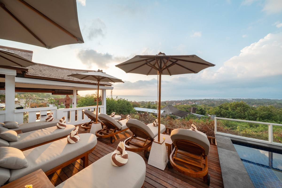 A spacious outdoor lounging area is equipped with multiple sunbeds and umbrellas. Views of the surrounding landscape are available, with a shimmering pool at one end. The wood decking offers a warm contrast to the neutral tones of the furniture.