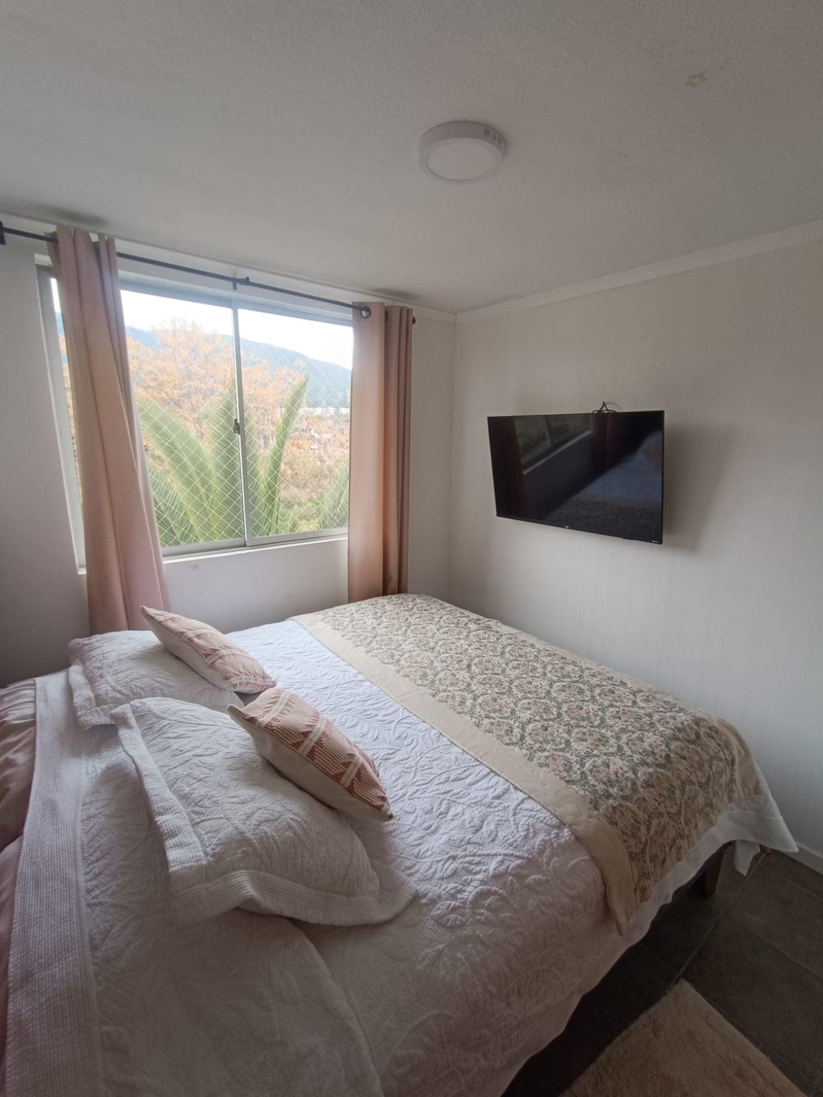 A bright bedroom features a spacious bed covered with a textured white blanket and decorative pillows. A flat-screen television is mounted on the wall opposite a large window, allowing natural light to fill the room and offering views of the surrounding greenery.