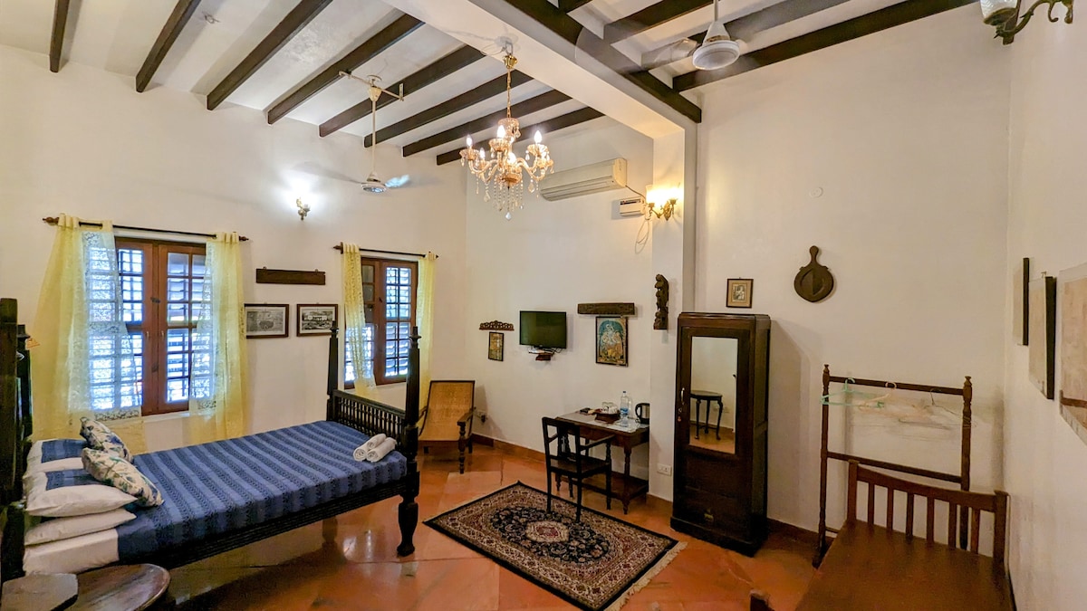 A spacious room features a double bed with a striped blue coverlet. Vintage furniture, including a wardrobe and writing desk, is present. Natural light enters through two windowed sections adorned with light curtains, and the ceiling is accentuated by wooden beams and a chandelier.