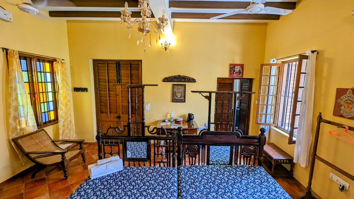 The twin bed room features wooden furniture and a colorful patterned bedspread. Natural light enters through multiple windows with light curtains, illuminating the warm yellow walls. A seating area includes a traditional chair and a small table with a kettle, while a light fixture adds additional charm.
