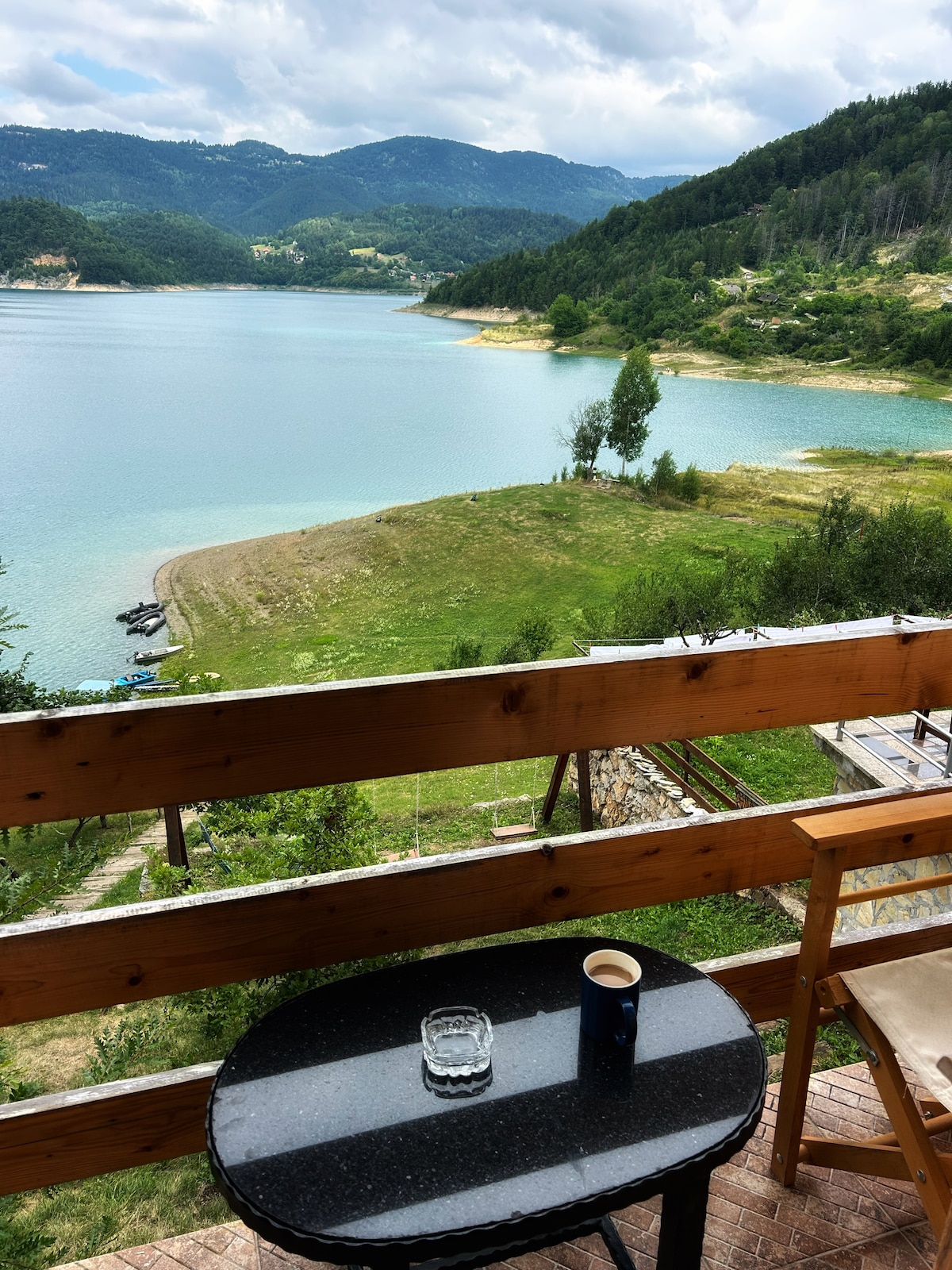 A balcony overlooking Lake Zaovine is depicted, featuring a small black table with a glass and a cup. The serene water reflects the surrounding green hills and mountains under a partly cloudy sky, creating a calming view for guests.