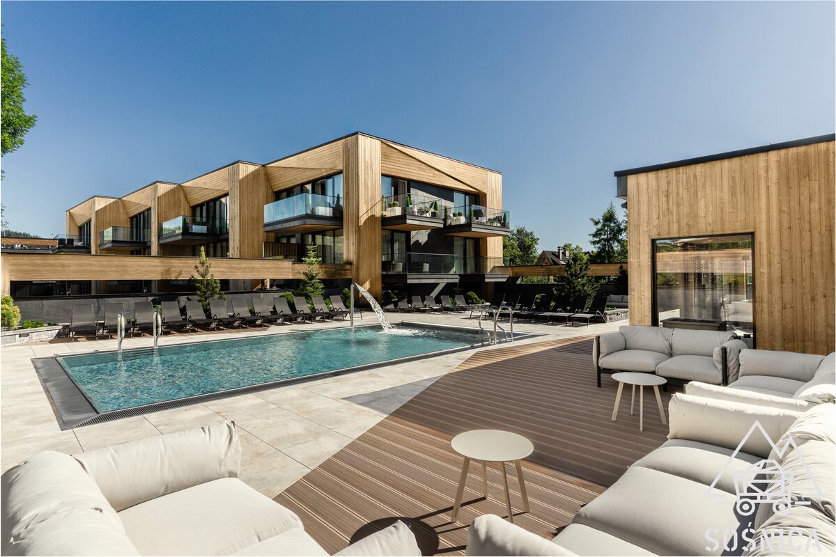 An outdoor area features a heated swimming pool surrounded by wooden decking and lounge chairs. Modern wooden architecture is visible in the background, with expansive glass windows reflecting the blue sky. Comfortable seating is arranged nearby, creating a relaxing environment.