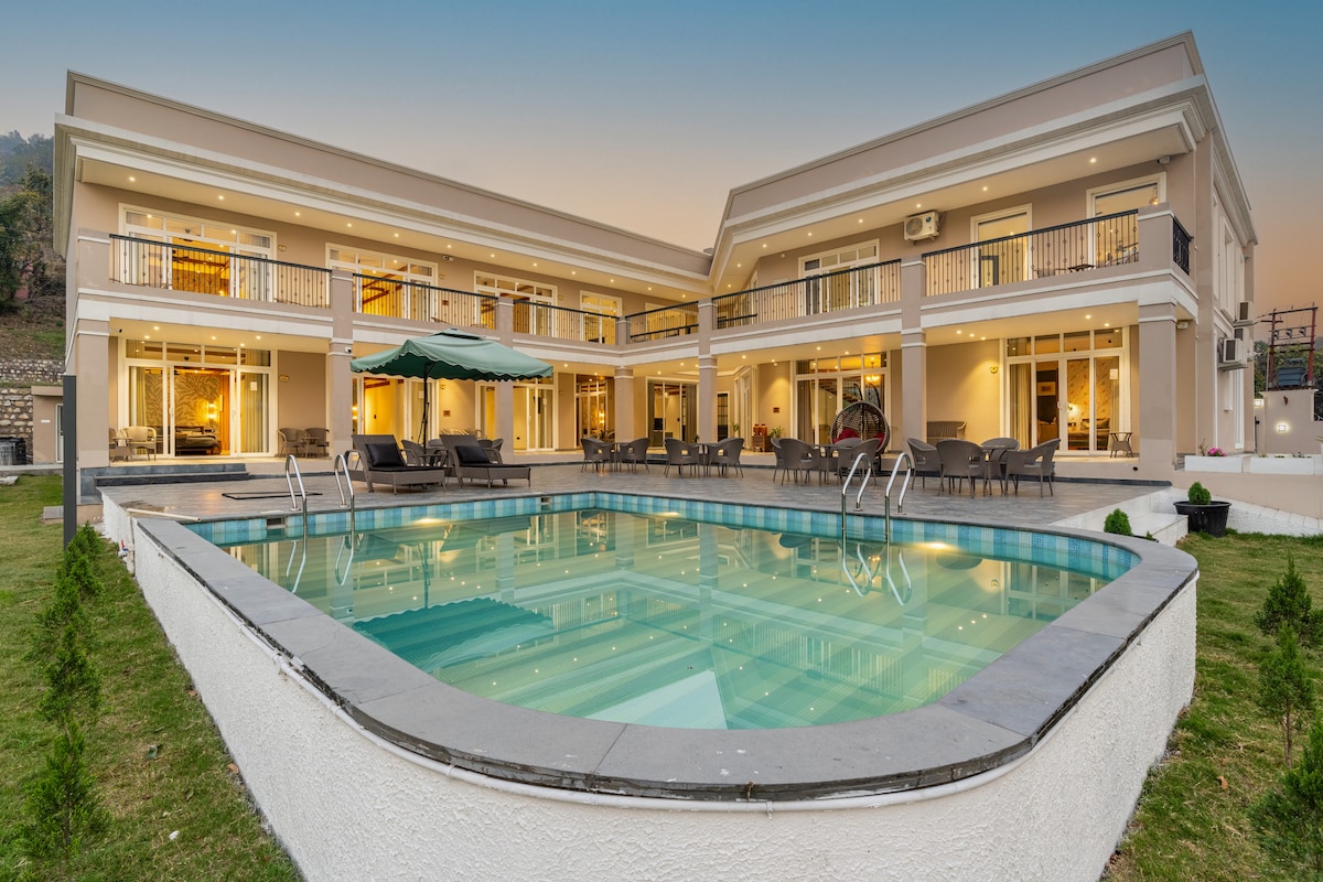 The villa's exterior features a spacious layout with a private pool at the center, surrounded by comfortable lounge seating. Large glass doors and windows provide views into the well-lit living spaces, inviting relaxation. The tranquil atmosphere is enhanced by greenery and the evening sky.