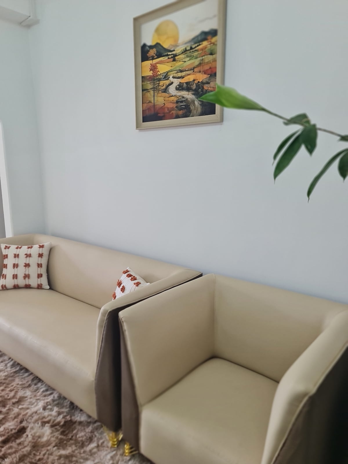 A light-colored sofa and matching armchair are showcased against a neutral wall. Decorative cushions add a touch of color, while a piece of artwork hangs above, featuring an abstract landscape. A green plant is positioned nearby, contributing to the overall fresh ambiance.