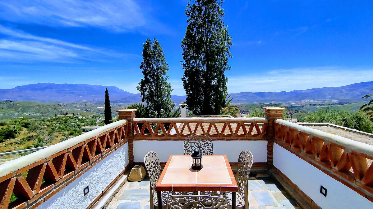 A spacious terrace offers unobstructed views of the surrounding landscape, featuring a tiled table with intricate metal chairs. Lush greenery and distant mountains are visible under a blue sky, enhancing the peaceful environment.