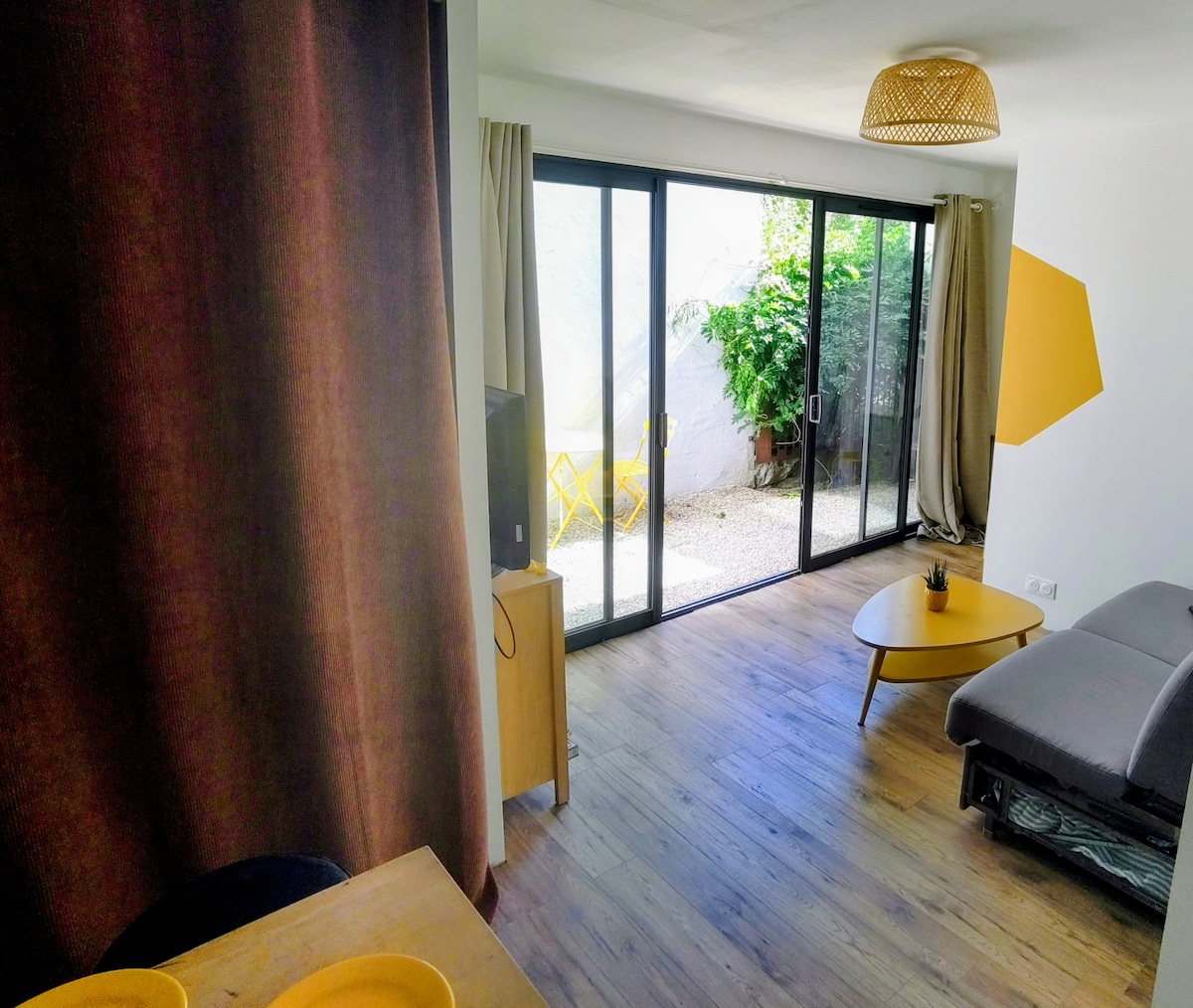 An inviting living area features sliding glass doors leading to a small outdoor space. Minimalistic furniture includes a grey sofa and a round coffee table. Natural light fills the room, with dark curtains partially drawn and a vibrant yellow accent wall adding a touch of color.