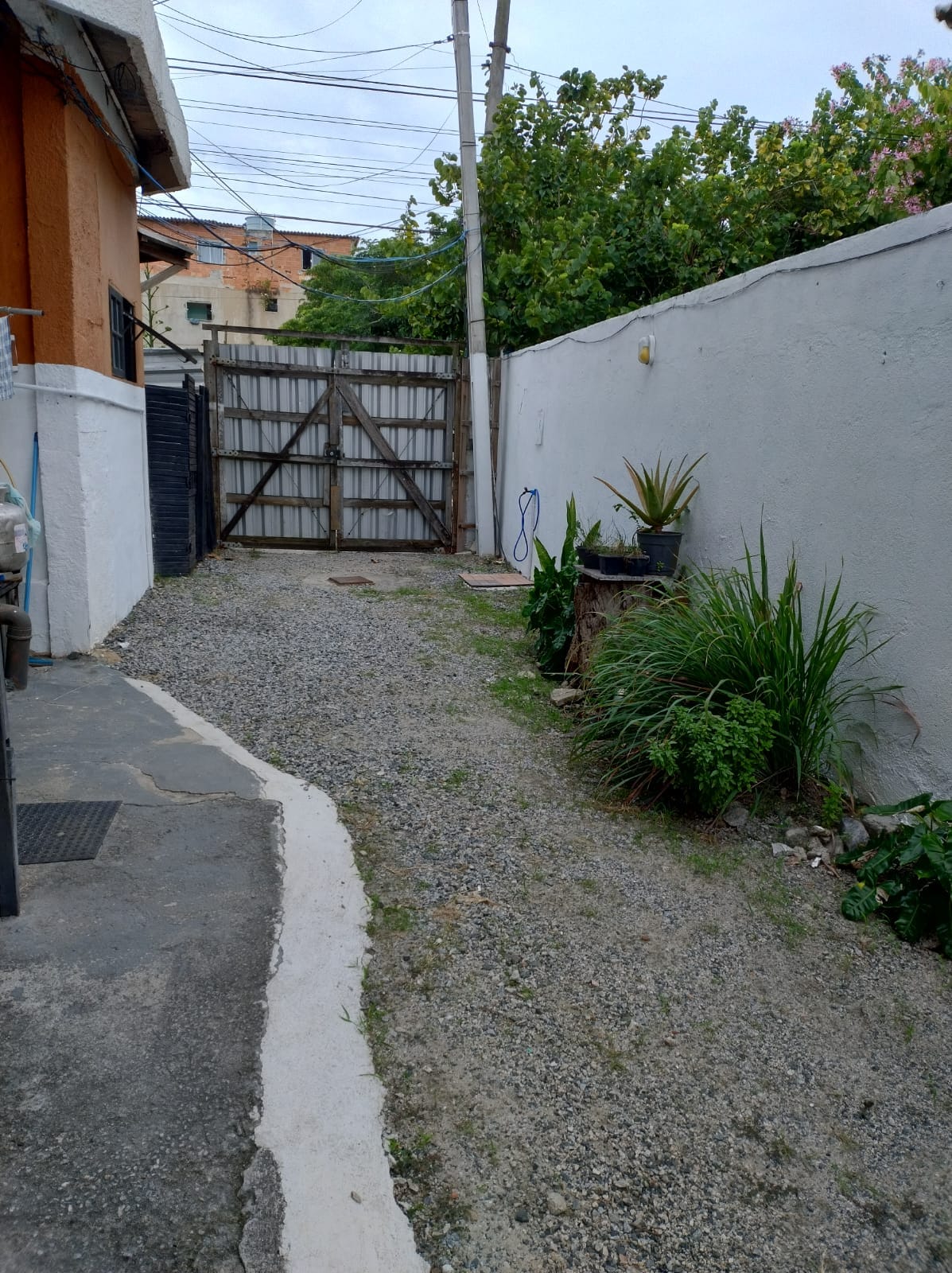 A gravel pathway leads toward a wooden gate, framed by a white wall on one side and greenery on the other. Various plants are visible along the edge, contributing to a natural ambiance. The area is quiet and appears well-maintained, providing access to additional spaces.