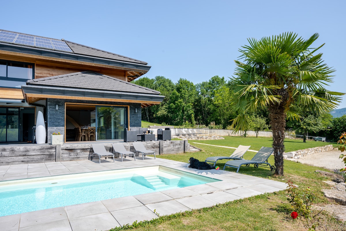 The image presents a modern two-story house featuring large windows and a spacious outdoor area. A covered pool is situated in the foreground, surrounded by lounge chairs. Lush greenery and a palm tree frame the property, enhancing the serene atmosphere.