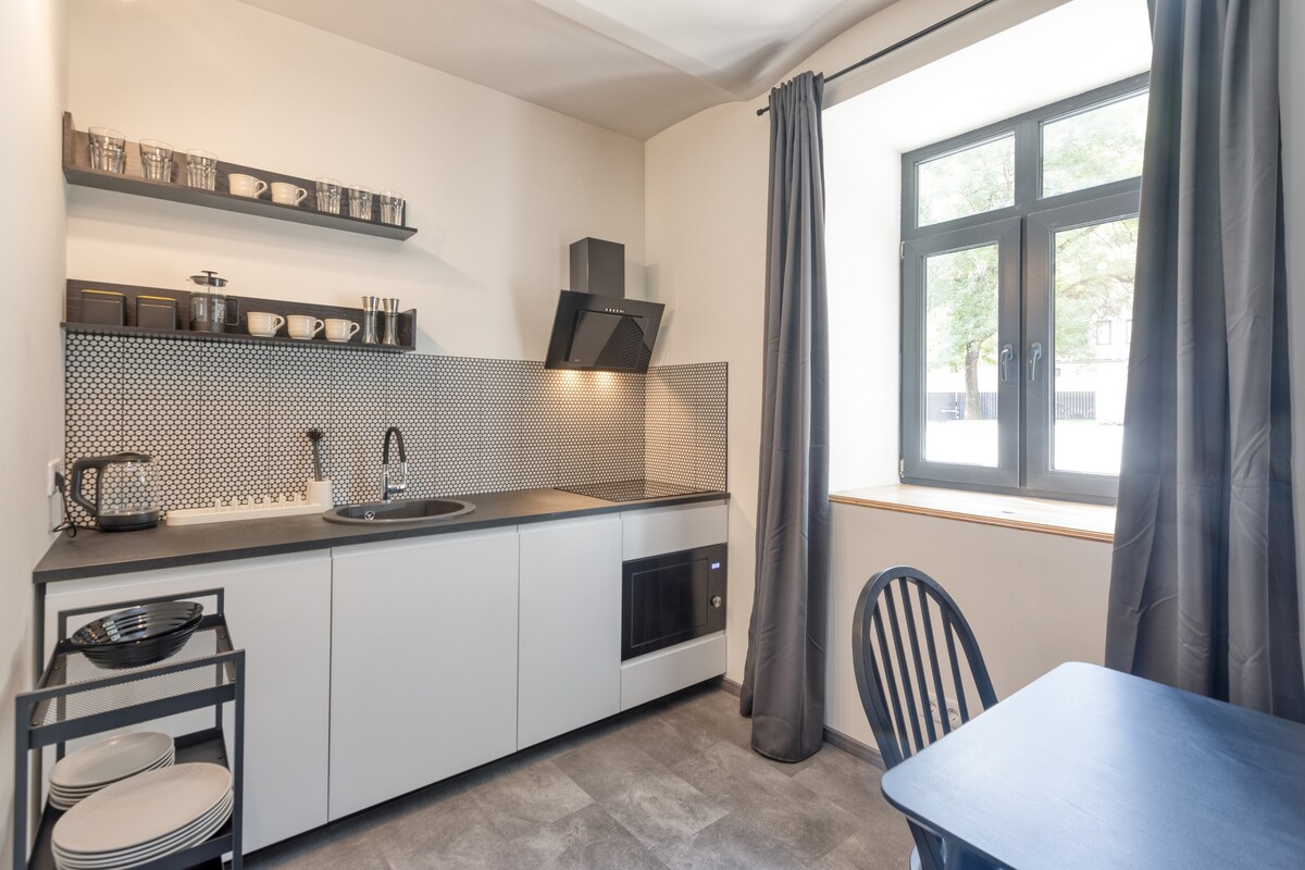 A modern kitchen is visible, featuring sleek cabinet storage and open shelving with dishware. A black countertop complements the grey walls and floor. A window allows natural light to enter the space, and a small dining table is situated nearby.
