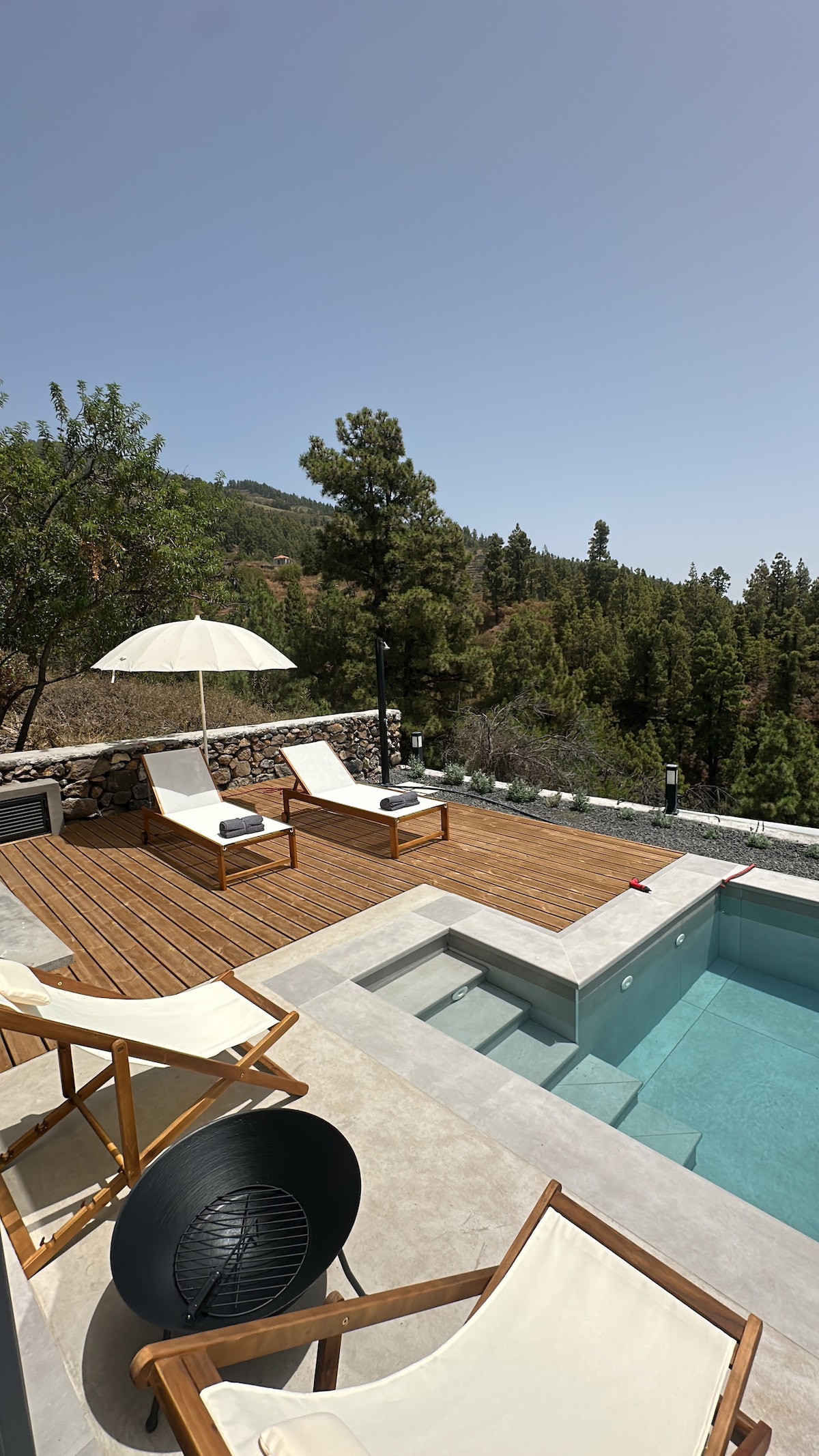 A serene outdoor space features a modern pool with clear water surrounded by a wooden deck. Two lounge chairs with cushions and a large umbrella offer shade, while a charcoal grill sits nearby against a backdrop of greenery and mountain views.