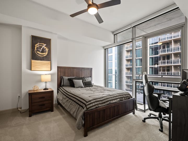 --Bračni krevet s modernim okvirom i uzglavljem od tamnog drva
– mekani neutralni posteljina s ukrasnim jastucima
– noćni ormarić sa svjetiljkom i zlatnim ukrasom
– apstraktna umjetnost na zidu koja dodaje dašak stila
– prozori od poda do stropa s izlazom na balkon