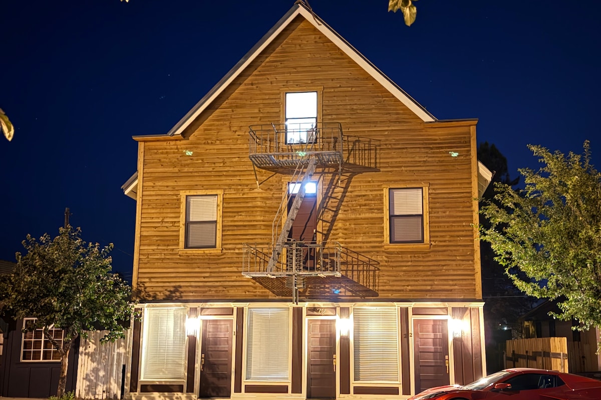 A three-story wooden building appears under a night sky, showcasing a fire escape on the front. Soft lighting from the windows and doorways highlights the rustic design. Surrounding greenery contributes to the welcoming atmosphere of the exterior.