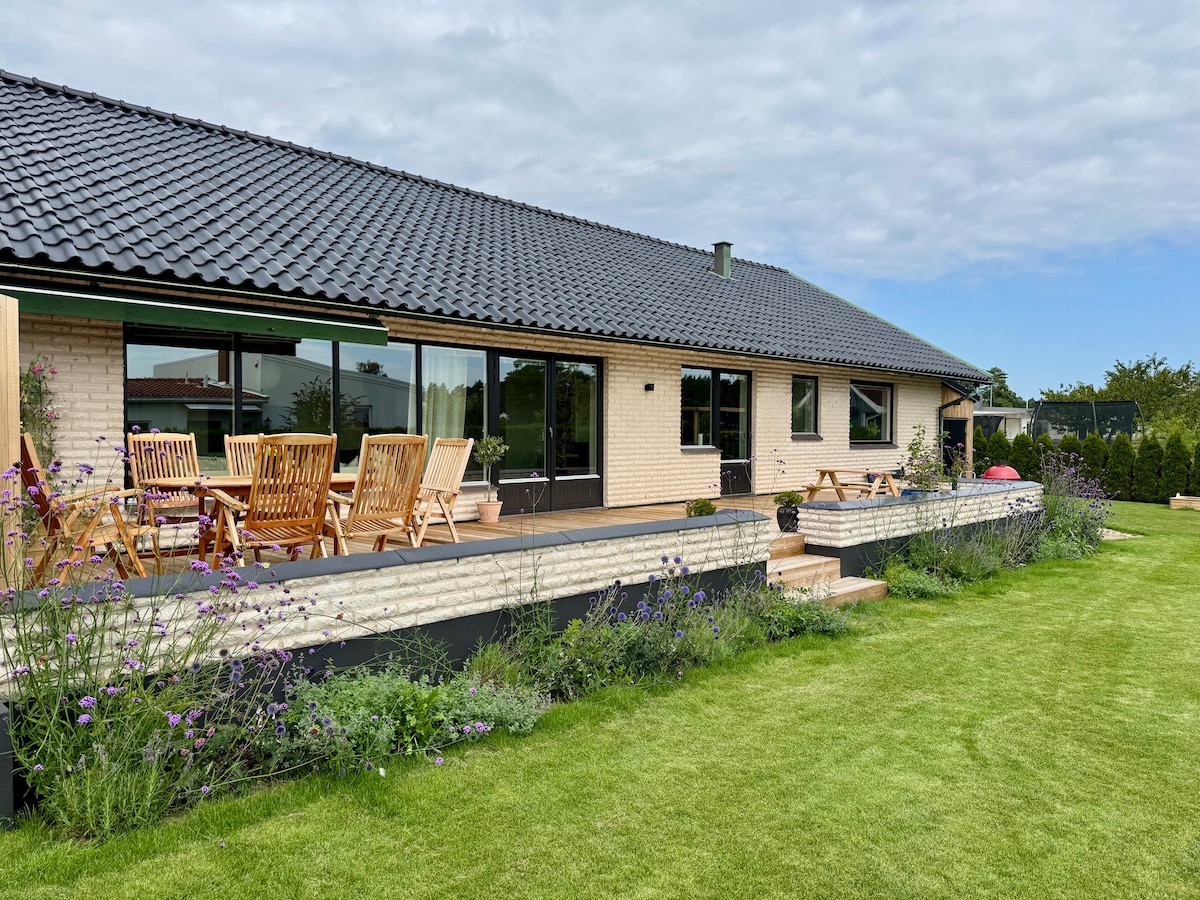 The exterior of a single-story house showcases a spacious wooden deck equipped with a dining area for ten. Lush green grass surrounds the property, complemented by flowering plants along the deck's edge. Large windows allow natural light to enter, enhancing the outdoor views.