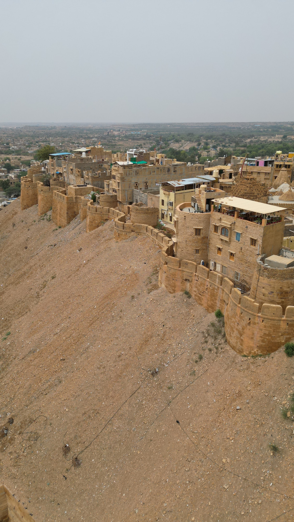 A panoramic view of the fort showcases fortifications and stone buildings perched on a rocky hillside. The landscape features a dry, sandy terrain extending into the distance, while the architecture reflects traditional styles, highlighting the rich history of the area.