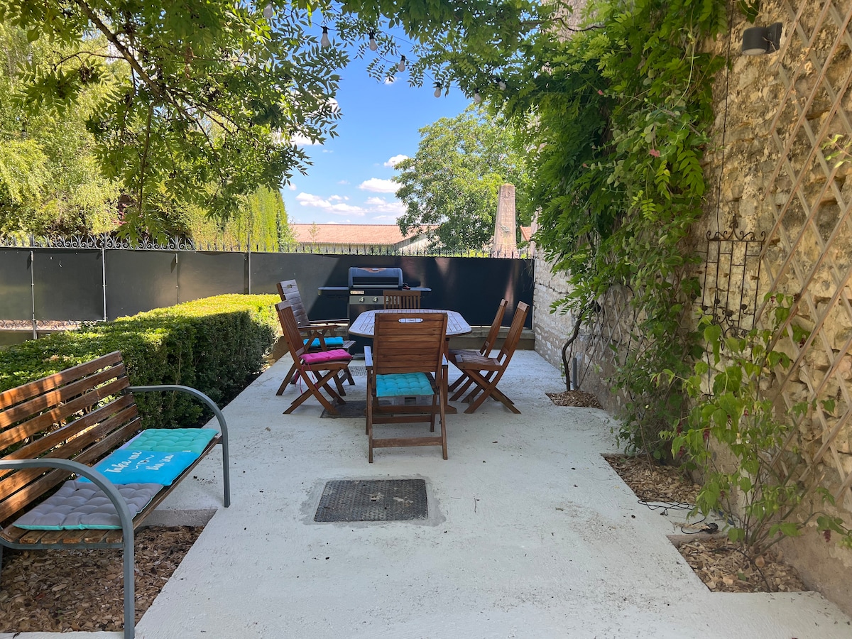 An outdoor patio area is visible, featuring a wooden dining table surrounded by six chairs. A gas barbecue is placed nearby, and greenery lines the perimeter, offering shade. The space is paved with a light-colored surface, creating a bright and inviting atmosphere for outdoor dining.