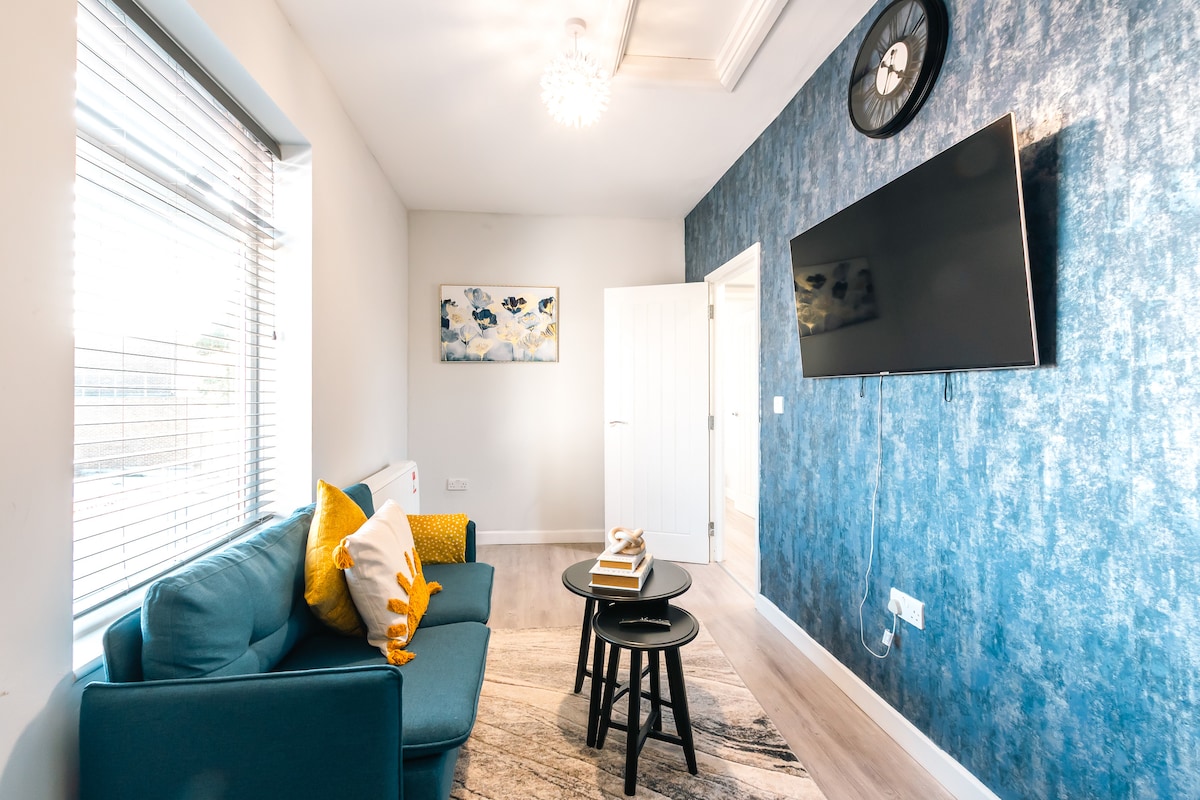 A cozy lounge area is presented with a teal sofa adorned with yellow accent pillows. A small round table holds a stack of books. A large wall-mounted TV is visible opposite the sofa. Natural light filters through the window, enhanced by light-colored blinds.