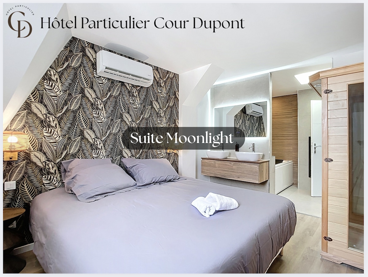 A cozy bedroom features a spacious bed dressed with soft grey linens. An accent wall adorned with tropical leaf patterns adds character. A pathway leads to a modern bathroom showcasing sleek fixtures, complemented by natural light from nearby windows and stylish wood elements throughout.