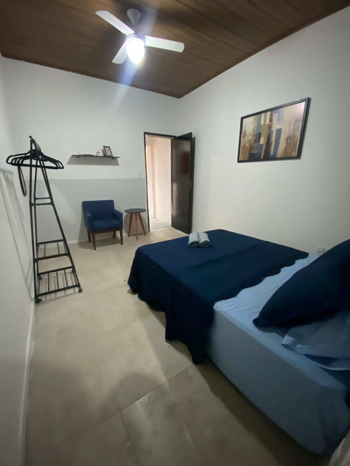 A simple and clean bedroom is depicted, featuring a spacious bed dressed in blue linens. A chair and small stool are positioned nearby, alongside a clothing rack against the wall. A framed artwork adorns the wall, and light is provided by a ceiling fan.