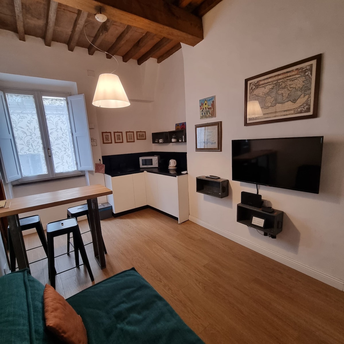 A dining area features a wooden table surrounded by black stools, positioned near a modern kitchen equipped with essential appliances. A wall-mounted television is visible opposite the seating area. Natural light enters through two windows with white shutters, showcasing the room's neutral decor.