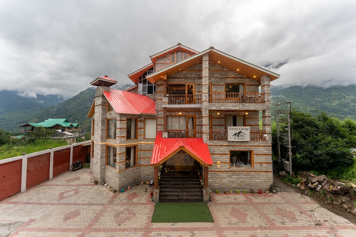 The exterior of the property features a multi-level stone building with wooden accents and a red roof. Surrounding greenery and mountains provide a scenic backdrop. A paved pathway leads to the entrance, while balconies and a terrace are visible on the upper levels.