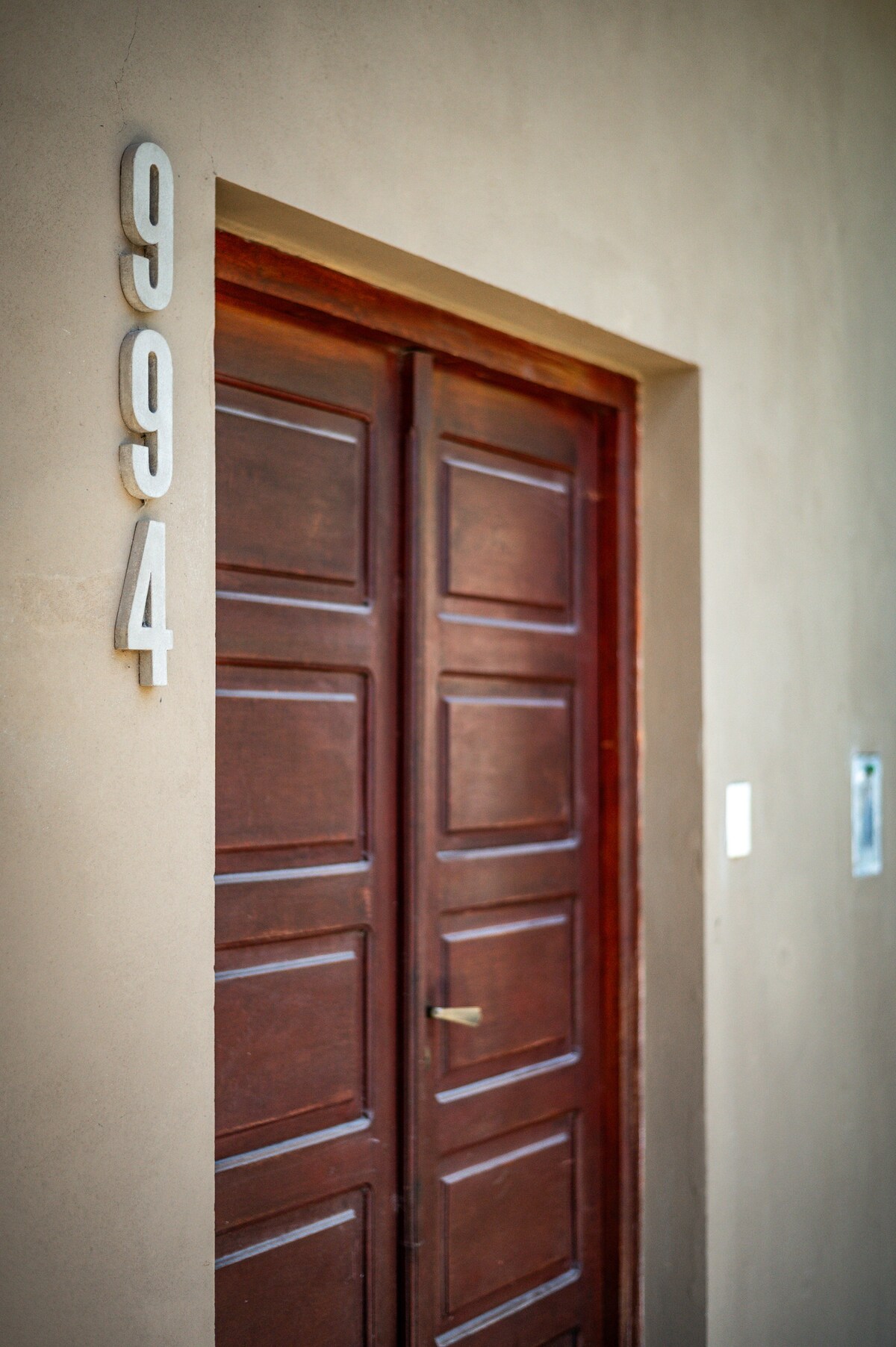 A wooden door with a rich brown finish is framed by a neutral-toned wall. The door features multiple panels and a simple handle, with house number 994 displayed prominently to the left in modern numerals. The entrance is well-lit, providing a welcoming appearance.