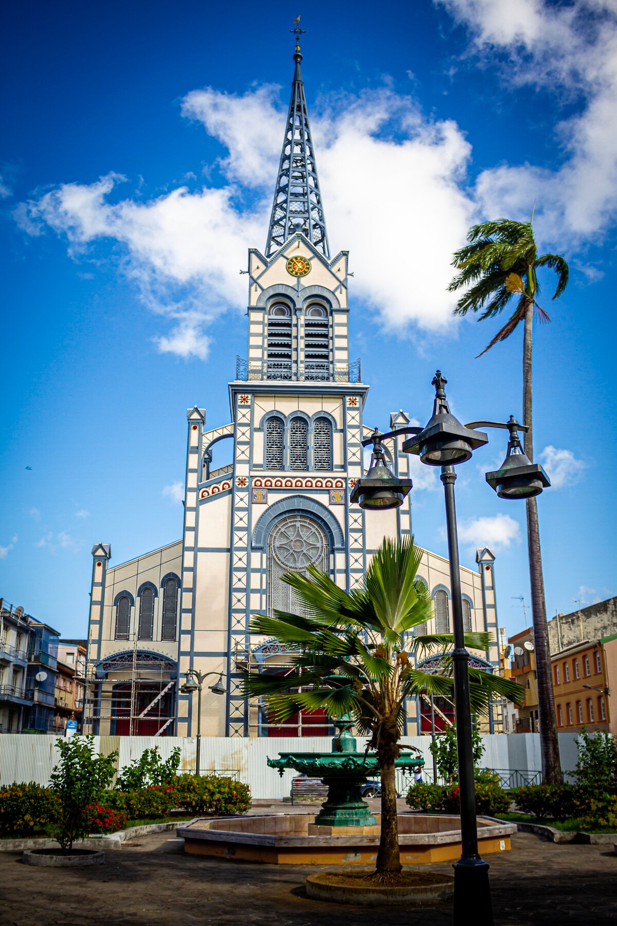 A striking church features a tall, ornate steeple and intricate architectural details. Surrounding greenery and a decorative fountain are visible in the foreground, with palms swaying gently nearby under a clear blue sky with scattered clouds.
