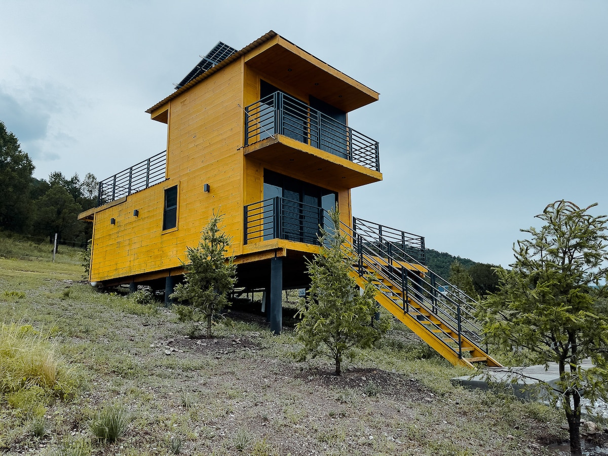 A modern two-story cabin constructed in yellow wood features large glass windows on each level. A wide staircase leads to the entrance, surrounded by gently sloping terrain and small evergreen trees. Solar panels are positioned on the roof, emphasizing a sustainable design.