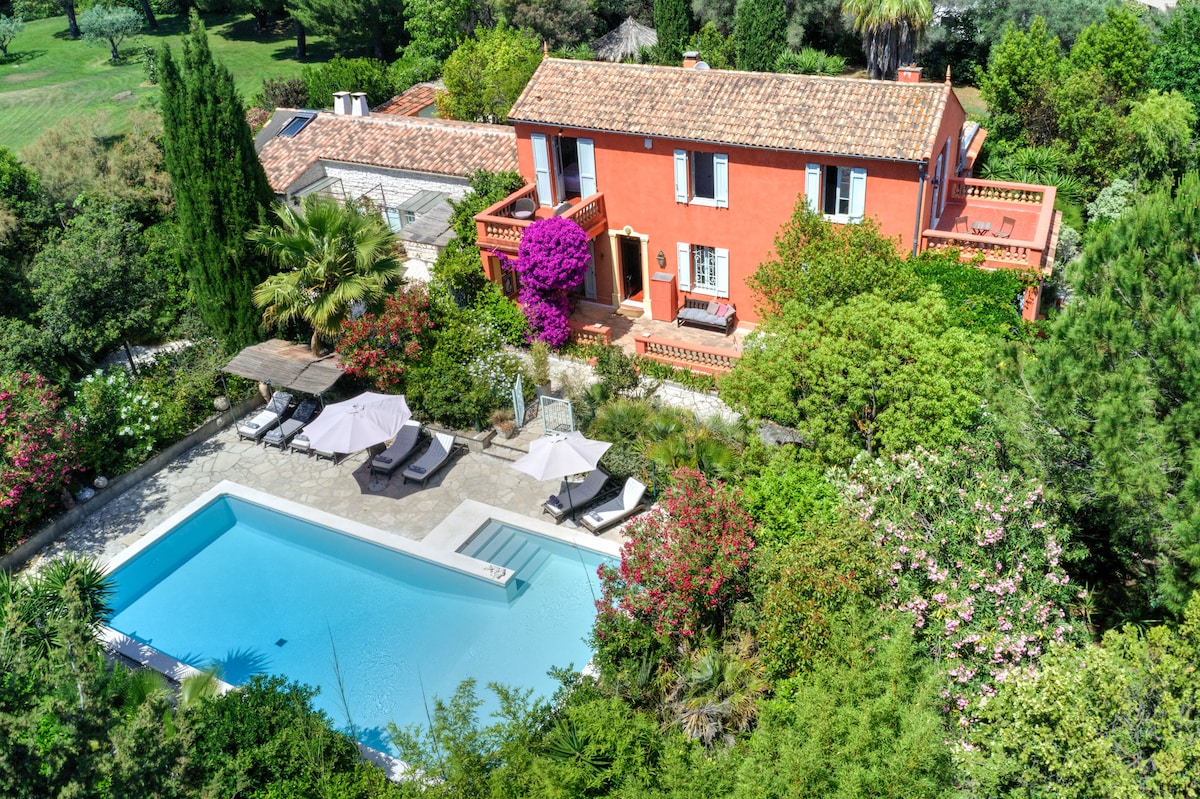 An aerial view reveals a vibrant property surrounded by lush greenery and colorful flowering plants. A clear pool is framed by sun loungers and umbrellas, while the house features terracotta walls and a tiled roof, with balconies overlooking the garden.