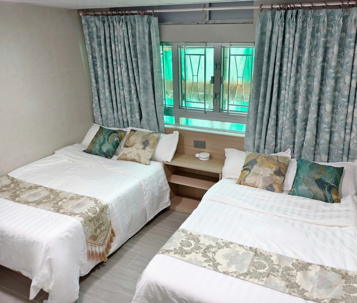 The room contains two spacious beds dressed in crisp white linens, with decorative pillows enhancing the aesthetic. A bedside table holds small plates, while light-filtering curtains partially cover the windows, creating a comfortable and well-lit atmosphere.