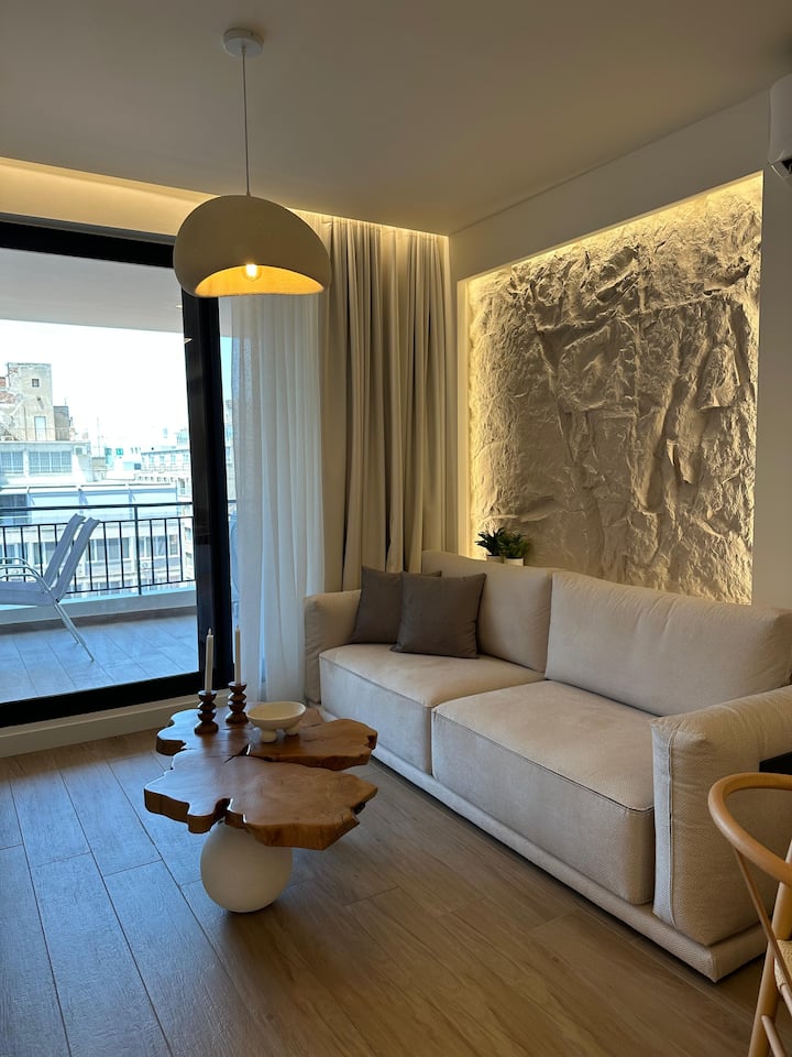 הסלון המודרני הזה כולל ספה בצבע קרם, שולחן קפה מעץ ייחודי, קיר אבן עם מרקם ותאורה אחורית חמה, ווילונות מהרצפה עד התקרה. גווני צבע טבעיים, תאורה רכה ועיצוב אלגנטי יוצרים אווירה מינימליסטית נעימה אך מסוגננת.